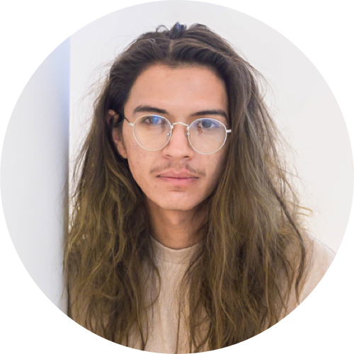 A close-cropped circular photographic headshot of a person with long, wavy hair and glasses wearing a beige shirt. They are looking directly at the camera and standing against a light background.